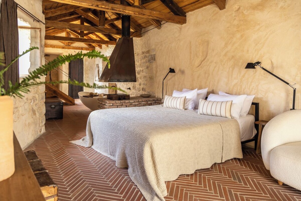 A cozy bedroom with a herringbone-patterned floor, a bed with white linens, a rustic stone wall, and a wooden ceiling.