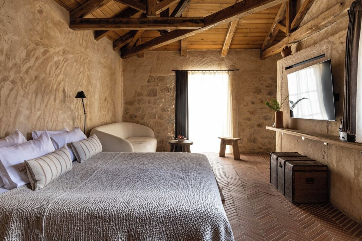 Un dormitorio rústico con una cama grande, techo de madera, paredes de piedra, un televisor y una mesita con una planta.