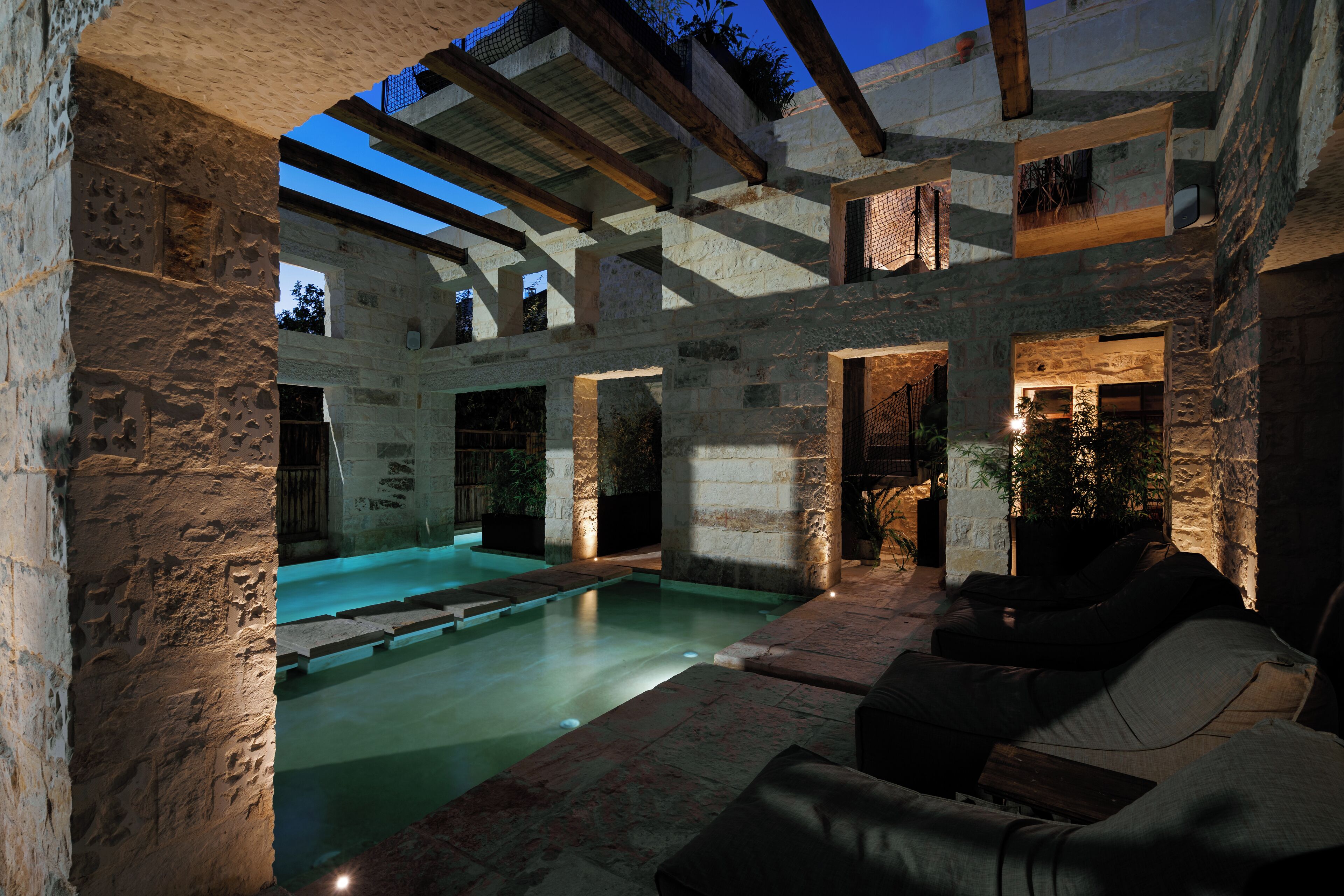 A stone-walled outdoor pool area with a wooden pergola overhead, stone seating, and a view of the sky.
