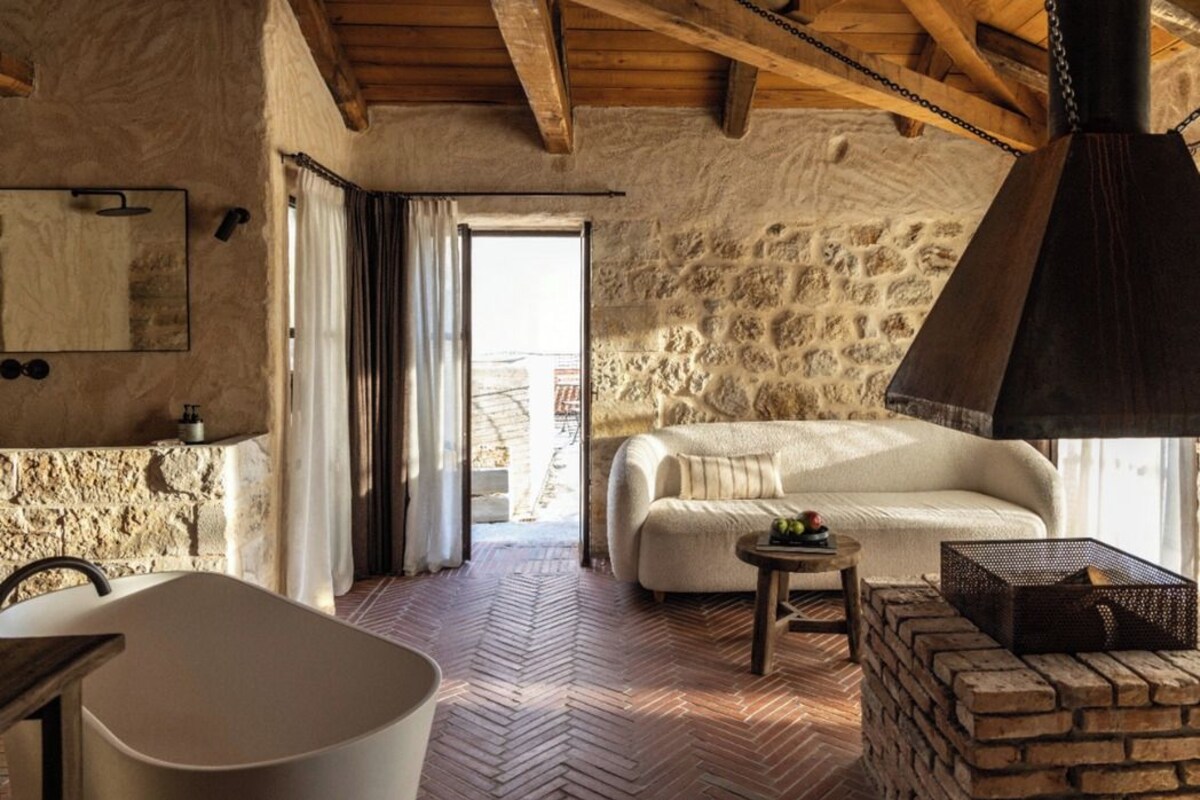 Una habitación rústica con pared de piedra, techo de madera, una bañera blanca, un sofá y una pequeña mesa con una cesta.