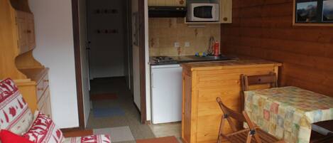 A compact kitchen with wooden cabinets, a microwave, and a small refrigerator. There is a dining area with a table and chairs, and a sofa with patterned cushions.