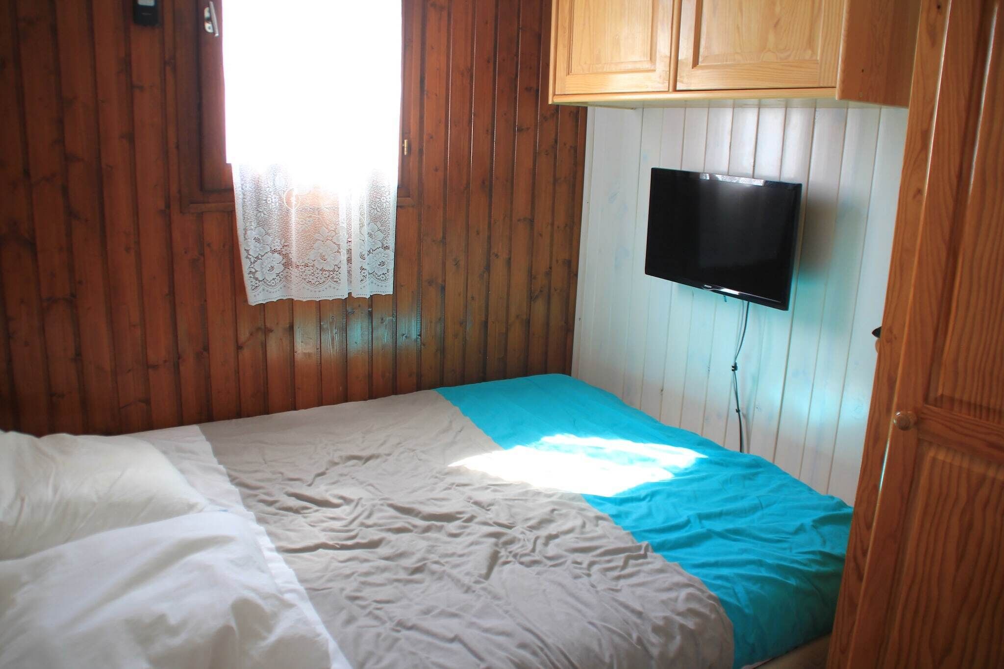Ein Zimmer mit Holzwänden, einem Bett mit blauer und weißer Bettdecke, einem an der Wand montierten Fernseher und einem Fenster mit Spitzenvorhang.