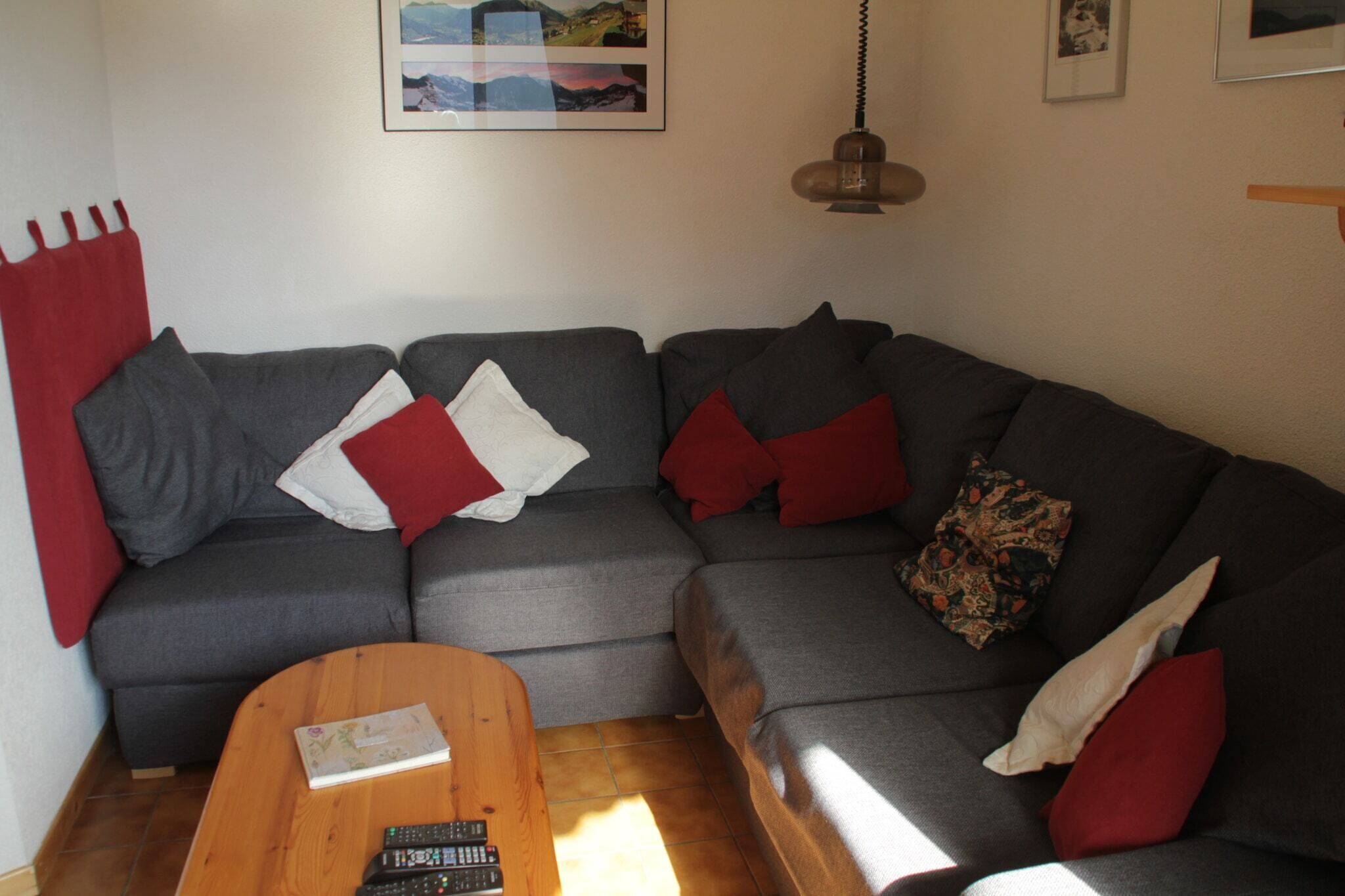 A modern living room with a grey sectional sofa, red and white cushions, a wooden coffee table, and a hanging pendant light.