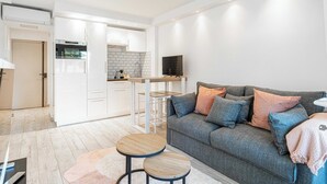 Un salon moderne avec un canapé gris, des tables rondes en bois et un coin cuisine doté de meubles blancs et d’un téléviseur.