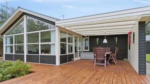 Et moderne hus med en træterrasse, et spiseområde med stole og et stort rum med vinduer.