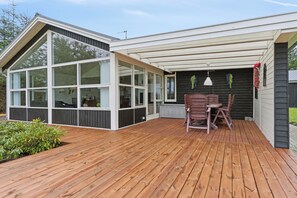 A modern house with a wooden deck, a dining area with chairs, and a large windowed room.