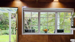 A room with a large window overlooking a garden, a wooden table with salt and pepper shakers, and a bowl of fruit on the windowsill.