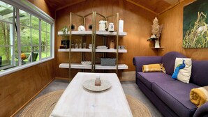 A cozy living room with a wooden interior, a purple sofa, a white coffee table, and a view of greenery through a large window.