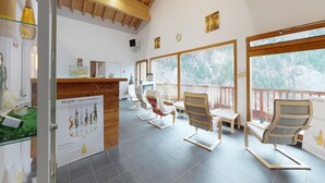 A spacious indoor area with wooden ceiling beams, large windows with a forest view, and several wooden chairs and tables. - Mountain view apartment in Orelle (Orelle)