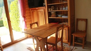 A wooden dining table with chairs, a TV on a shelf, and a view of greenery outside. - Apartment for 3 people with balcony (Châtel)
