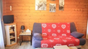Une pièce chaleureuse avec un mur en bois, un canapé recouvert d’une couverture à motifs de cœurs, une petite table avec une lampe et une étagère remplie de livres.