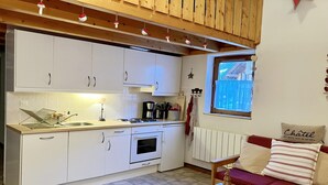 Une cuisine compacte avec des meubles blancs, un évier et une cuisinière. On y trouve une table en bois sur laquelle est posé un objet décoratif, devant un canapé rouge agrémenté d’un coussin sur lequel est inscrit le mot « Châtel ».