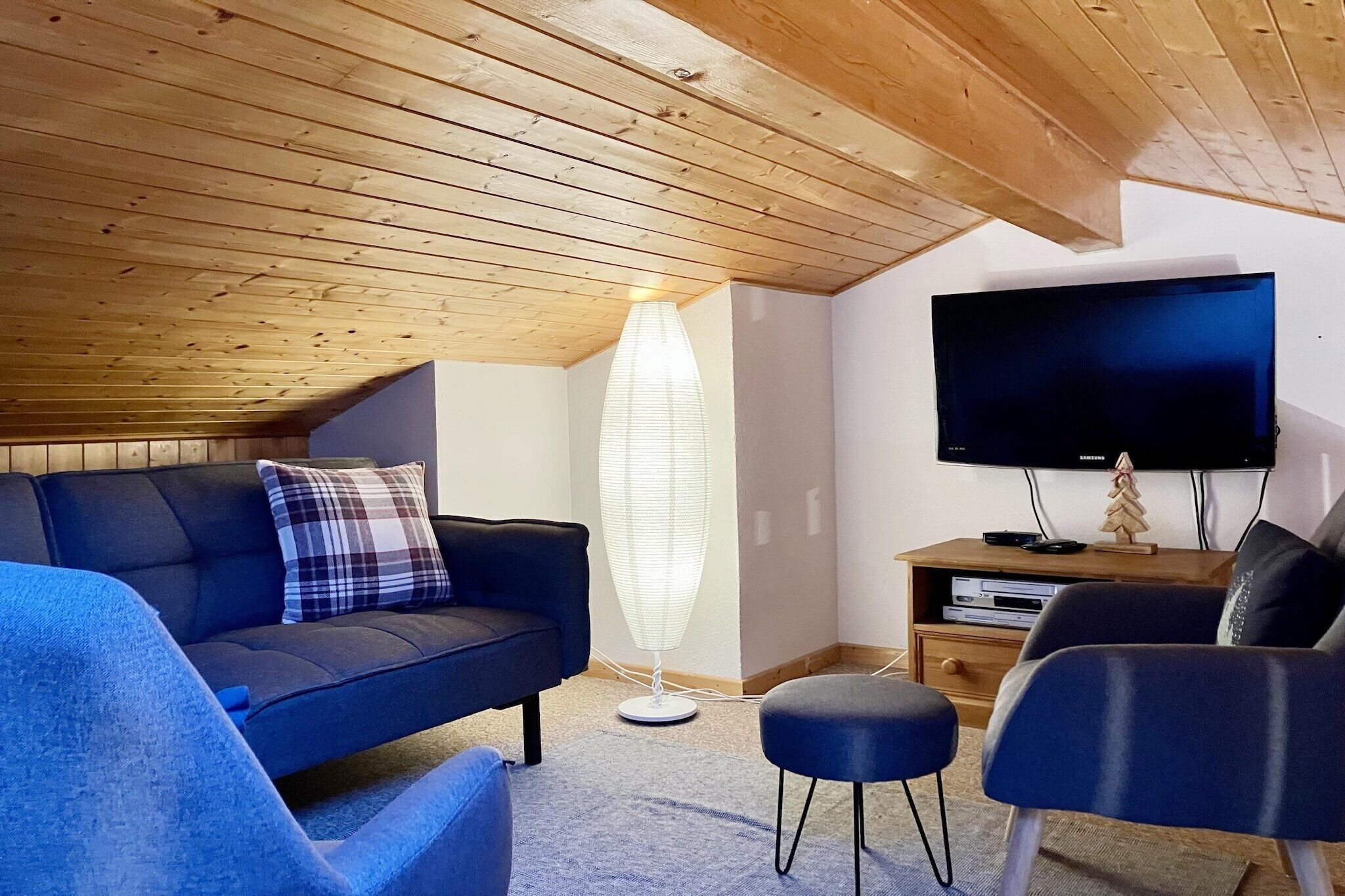 Un salon chaleureux avec un plafond en bois, un canapé bleu foncé, un lampadaire, un téléviseur à écran plat et un petit meuble en bois.
