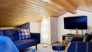 Un salon chaleureux avec un plafond en bois, un canapé bleu foncé, un lampadaire, un téléviseur à écran plat et un petit meuble en bois.