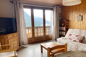 A cozy room with a wooden sofa, a television on a wooden cabinet, and a balcony with a view of mountains. - Apartment near centre Chatel with balcony (Châtel)