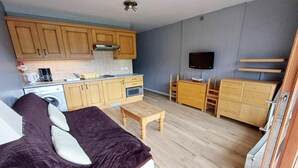 A compact living space with a kitchenette, a bed with a dark blanket, a small wooden table, and a TV mounted on the wall. - Apartment in Chatel for two people (Châtel)