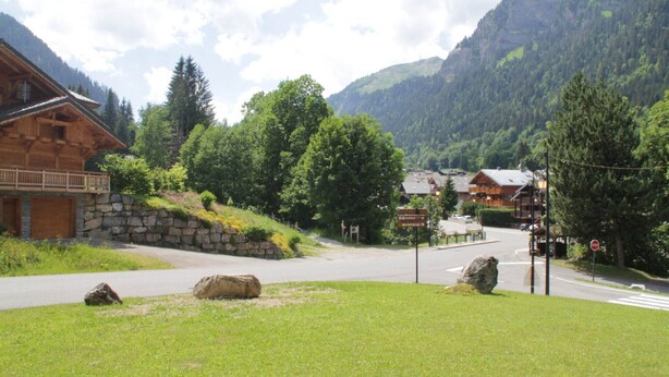 A mountain village with wooden buildings, a stone wall, and a road leading to a signpost.