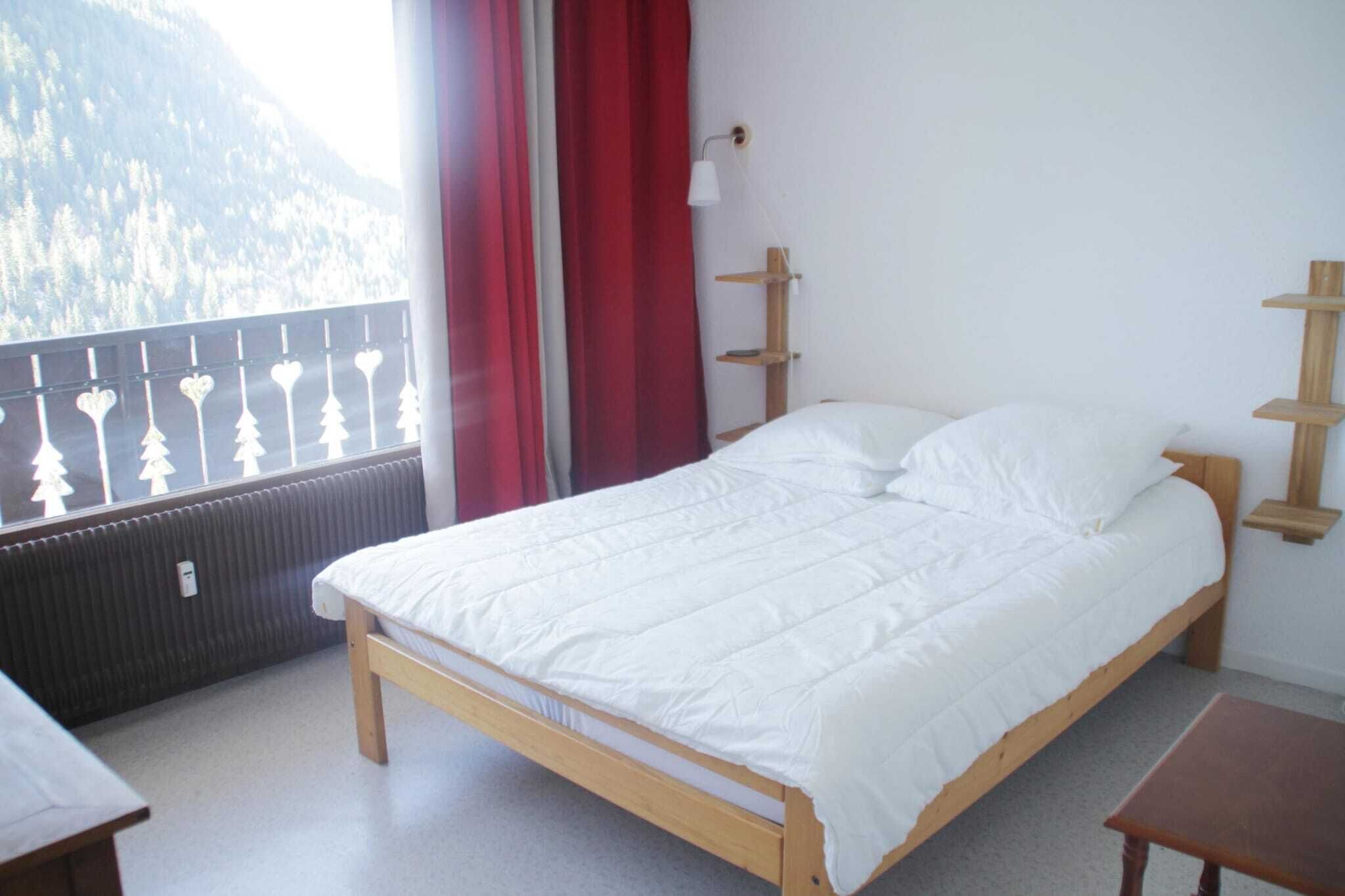 A single-bed room with a wooden frame, white bedding, a window with a view of a forested mountain, and a red curtain.
