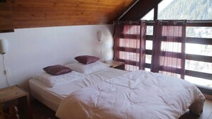 Un lit simple avec une literie blanche, dans une pièce aux murs en bois et dotée d’une fenêtre avec des rideaux.