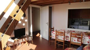 Une pièce chaleureuse avec un plafond en bois, un téléviseur posé sur un meuble en bois, une table à manger avec des chaises, et un coin cuisine visible à travers une porte ouverte.