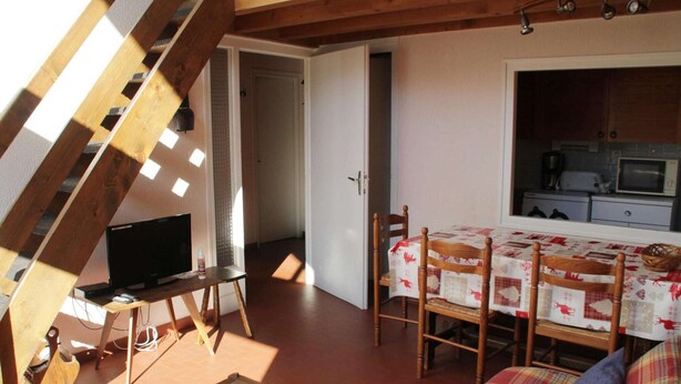 A cozy room with a wooden ceiling, a television on a wooden stand, a dining table with chairs, and a kitchen area visible through an open door.