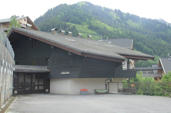 A building with a sloped roof, a sign reading "L'ANNA MARIA," and a view of a forested mountain in the background.