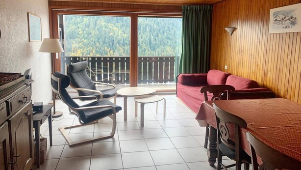 Una habitación acogedora con techo de madera, un sofá rojo, un sillón lounge negro, una mesita y vistas a un bosque a través de amplios ventanales.