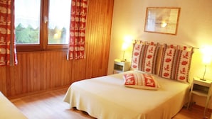 A bedroom with a wooden floor, a bed with a white bedspread, two bedside lamps, and red and white patterned curtains.