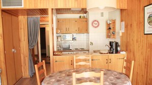 A compact kitchen with wooden cabinets, a microwave, and a kettle. There is a dining table with chairs and a clock on the wall.