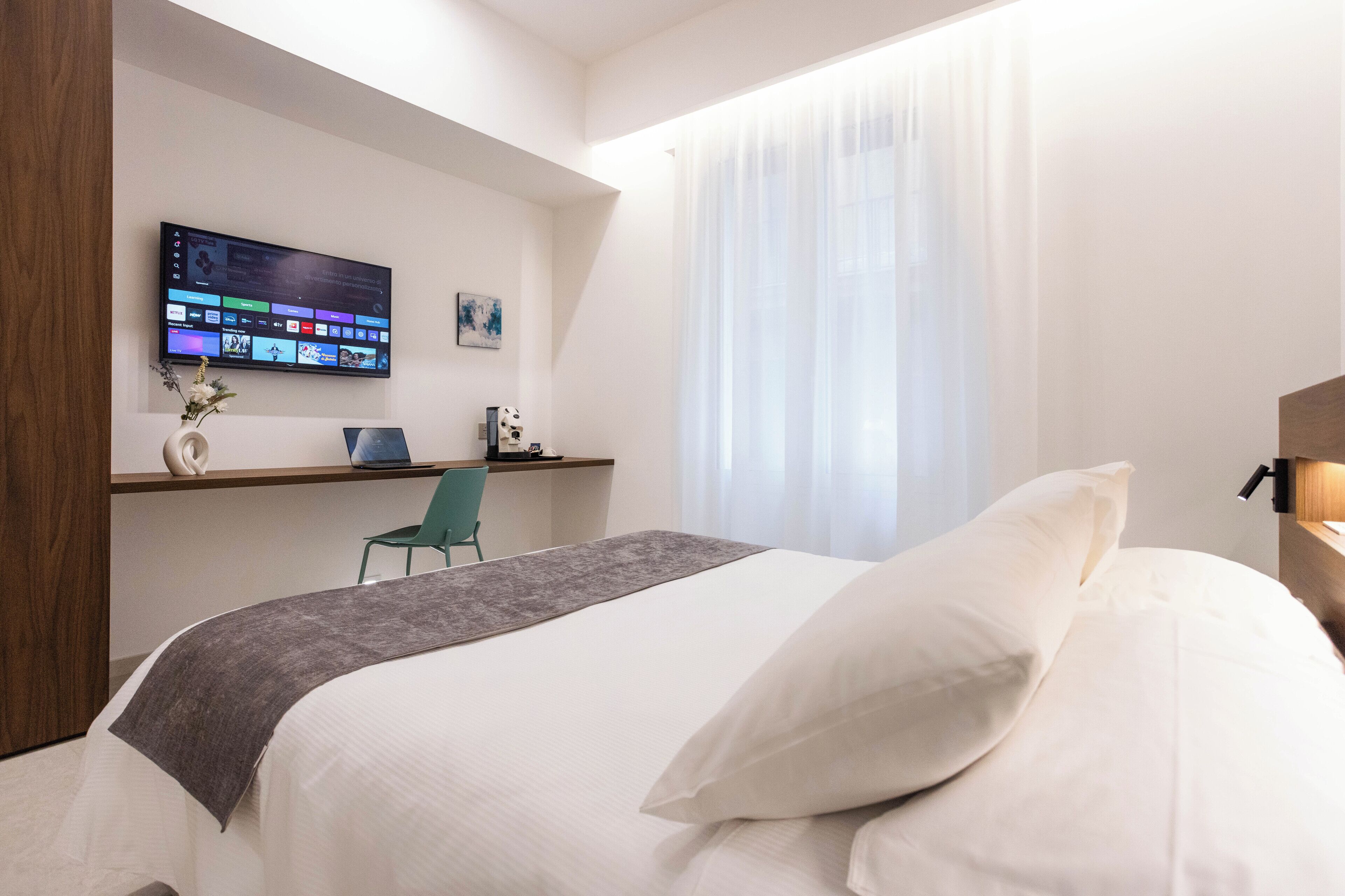 A modern hotel room with a bed, a desk with a laptop, a TV mounted on the wall, and a window with sheer curtains.