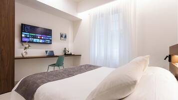 A modern hotel room with a bed, a desk with a laptop, a TV mounted on the wall, and a window with sheer curtains.