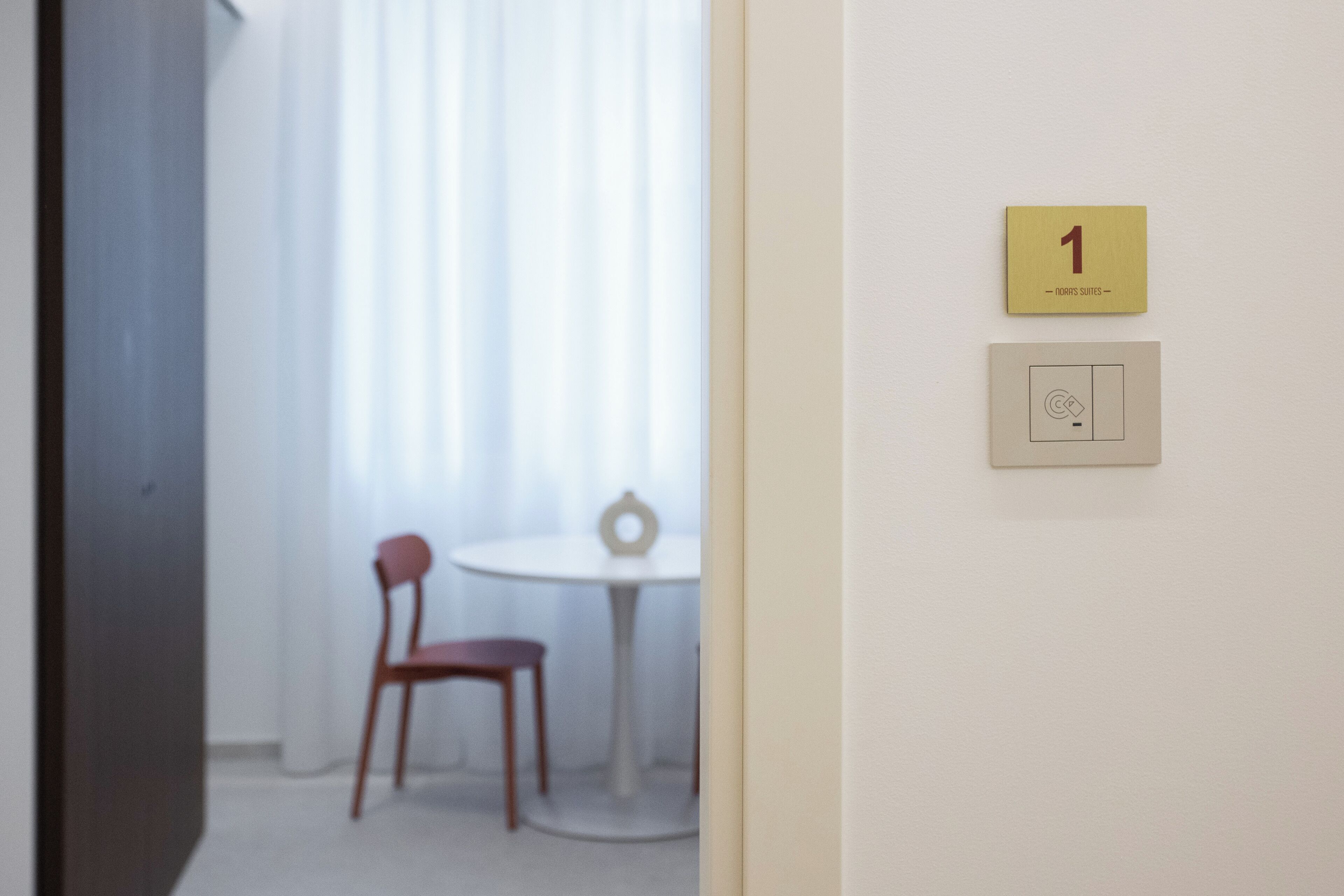 A room with a chair and a table, a door with a number sign, and a light switch on the wall.