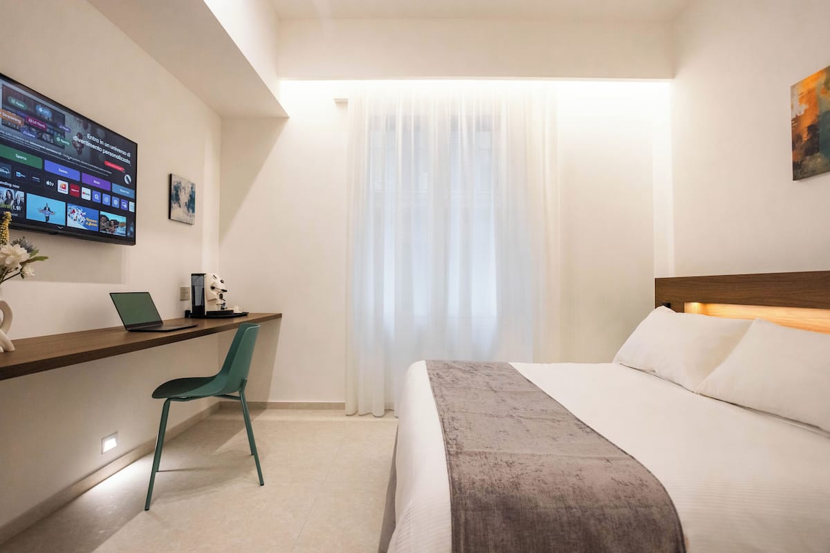 Quarto de hotel com uma cama, uma escrivaninha com um laptop, uma cadeira, uma TV fixada na parede e uma janela com cortinas leves.