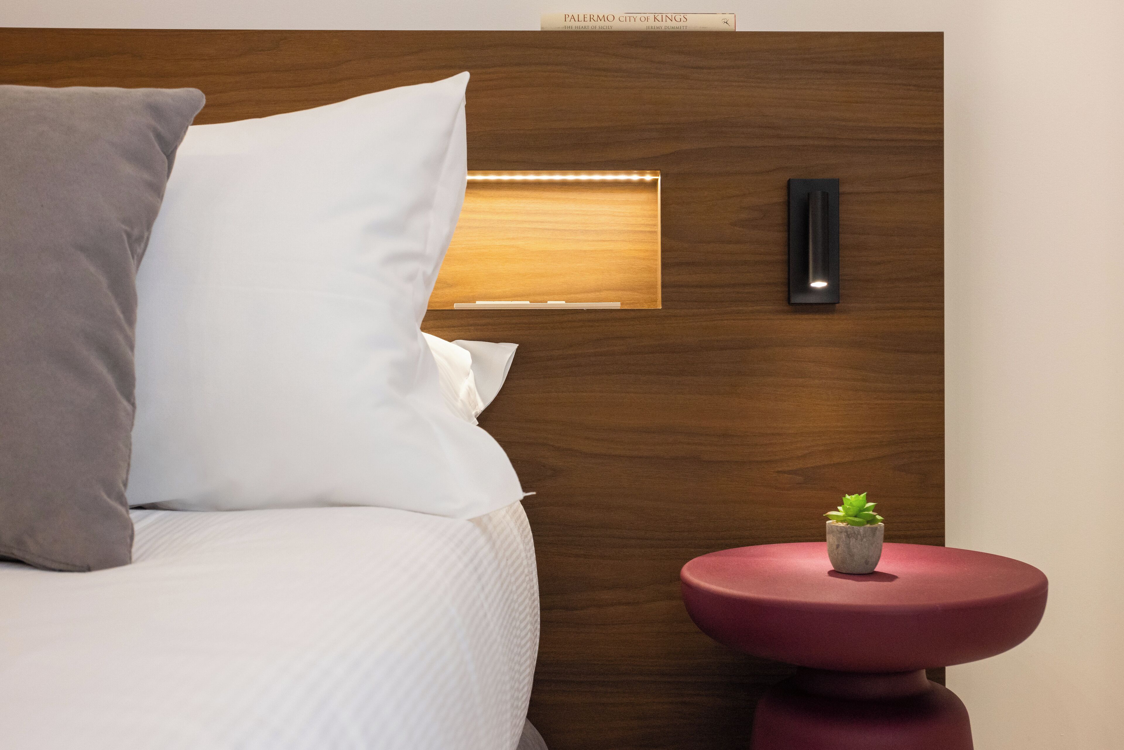 A neatly made bed with white and gray pillows, a small potted plant on a round red table, and a wooden headboard with a built-in light.