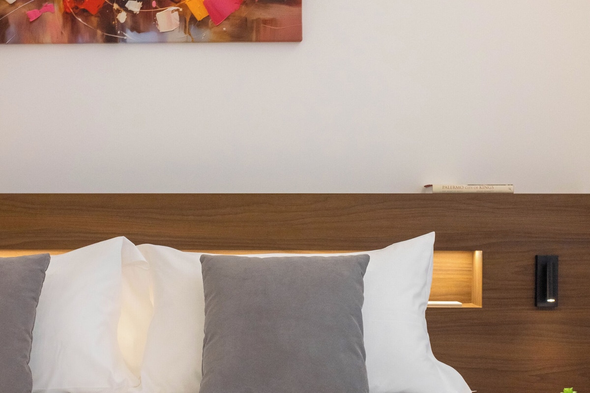 Uma cama bem arrumada com cabeceira de madeira, uma pequena mesa de cabeceira com uma planta, e um quadro abstrato colorido sobre a cama.