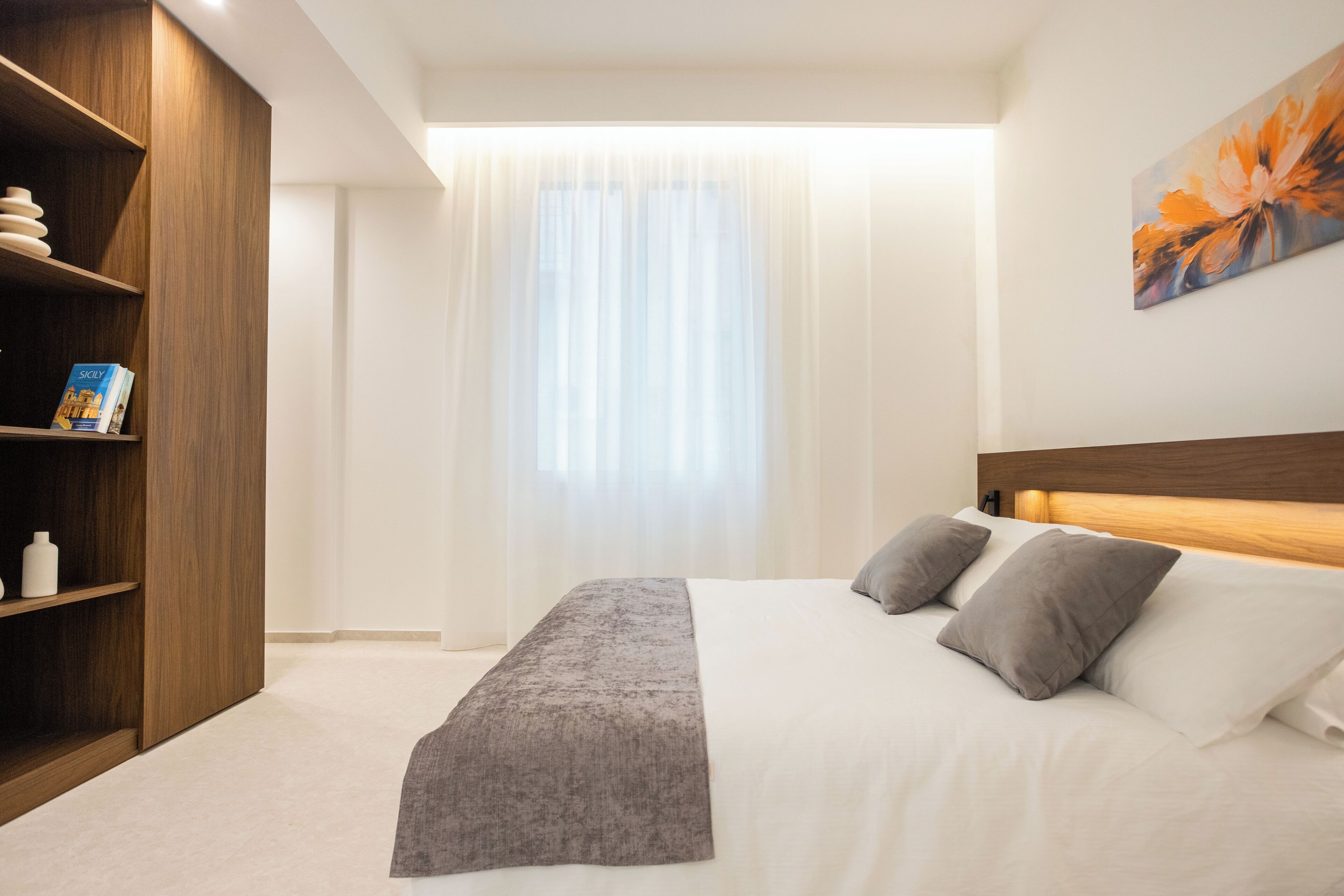 Une chambre à coucher comprenant un lit, une armoire en bois, une table de chevet, un tableau et une fenêtre avec des rideaux légers.