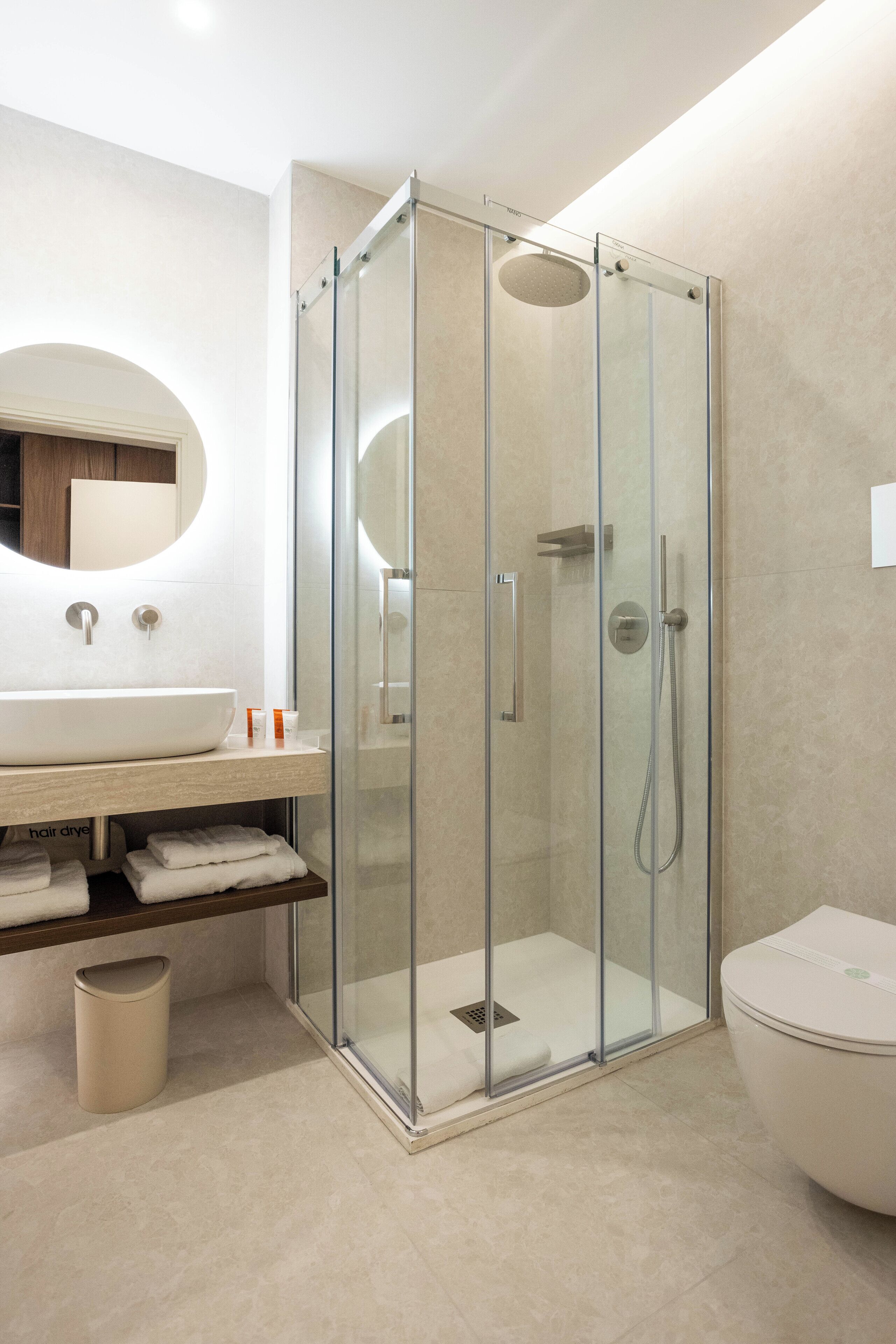 A modern bathroom with a glass-enclosed shower, a round mirror, and a white sink.