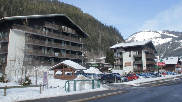 A multi-story hotel with balconies, a parking area, and a mountainous backdrop.