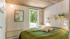 Un dormitorio con techo de madera, una cama con ropa de cama verde, una ventana con vista a la vegetación y una estantería con plantas.