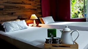 A hotel room with a wooden wall, a bed with white bedding, a nightstand with a lamp, and a window with a view of greenery. - Babblers Inn By GRB (Tinsukia)