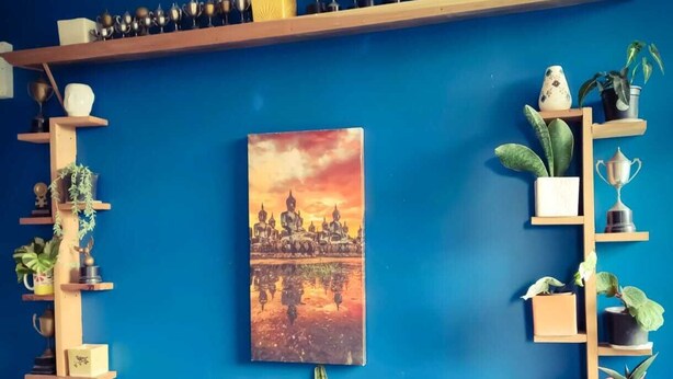 A room with a blue wall, a central painting of a temple, and shelves with trophies and plants. - Babblers Inn By GRB (Tinsukia)