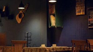 A dimly lit room with a guitar hanging on the wall, a table with chairs, and a bookshelf with books. - Babblers Inn By GRB (Tinsukia)