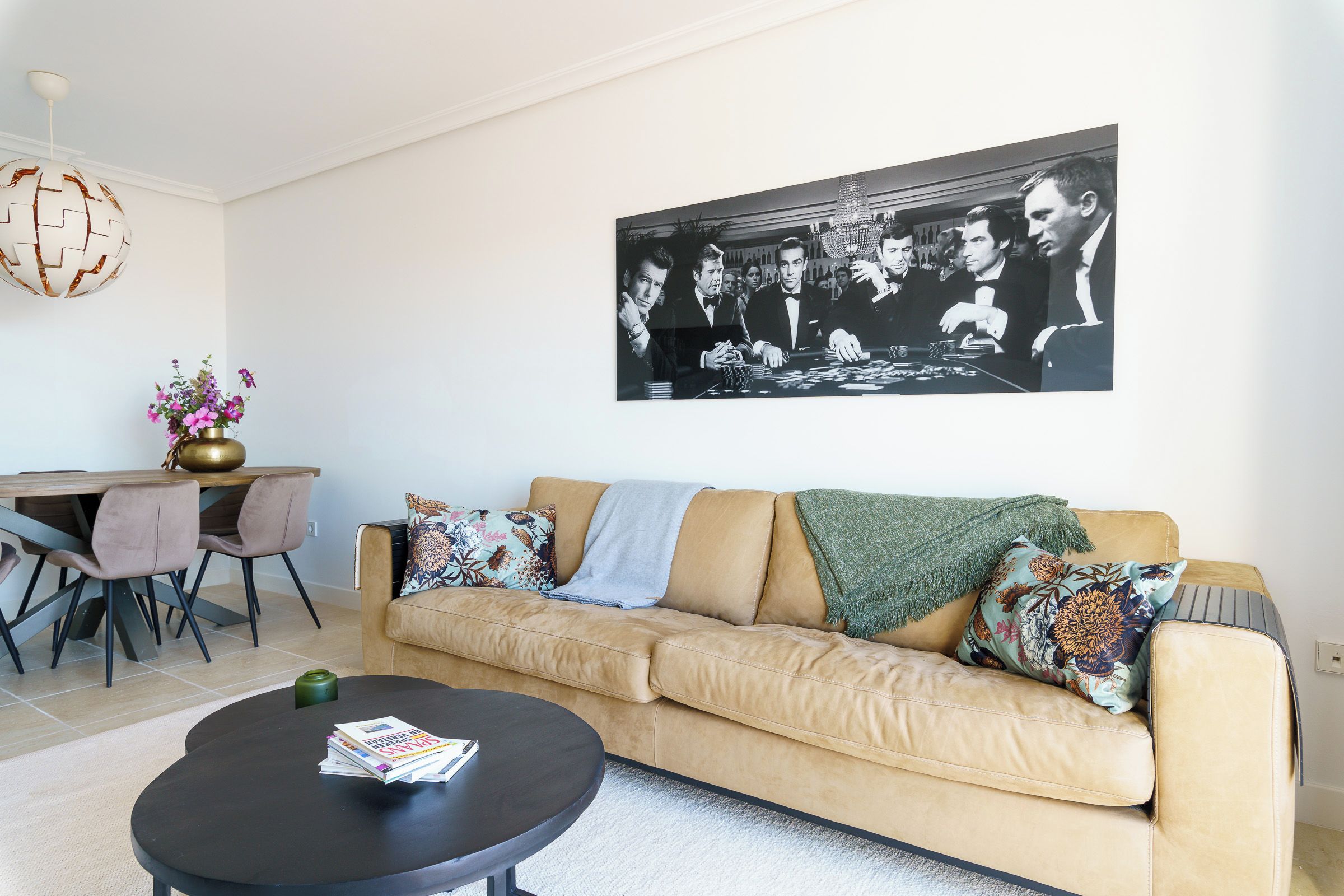 Una sala de estar con un sofá beige, una mesa de centro redonda, una zona de comedor con sillas y una lámpara globo fijada a la pared.