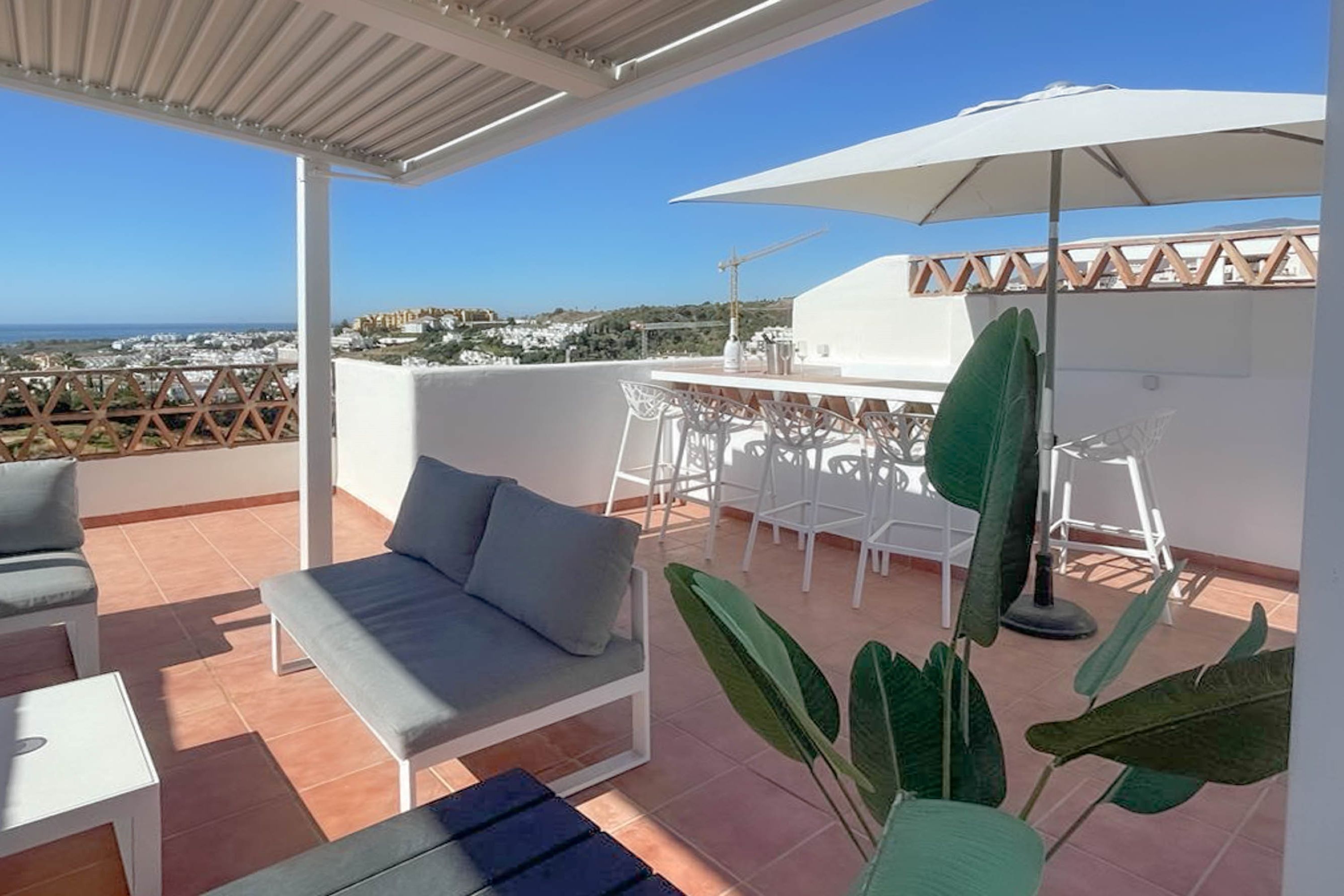 Eine Dachterrasse mit einem weißen Tisch und Stühlen, einer grauen Couch und einem großen Sonnenschirm.