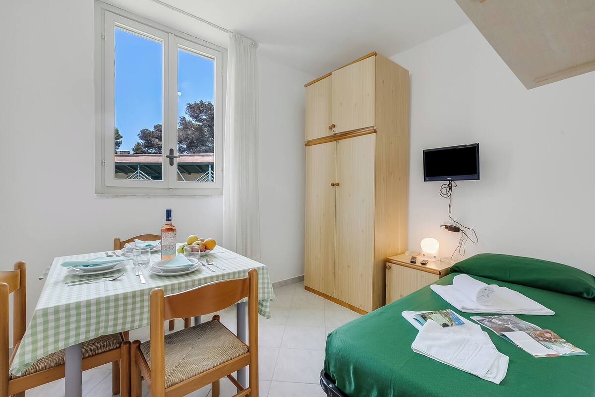 A room with a dining table set for two, a wardrobe, a bed with green bedding, a bedside table with a lamp, and a TV mounted on the wall.