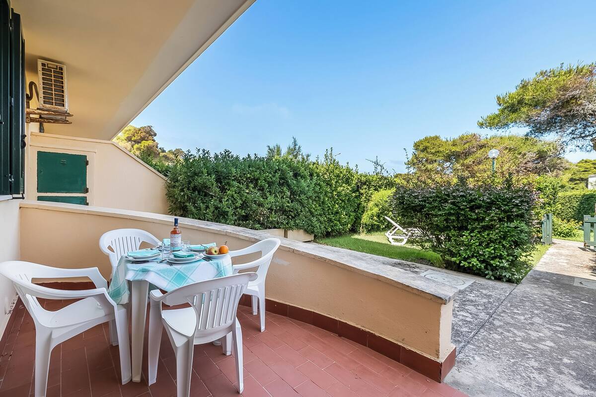 A balcony with a table set for dining, white plastic chairs, and a view of a well-maintained garden.