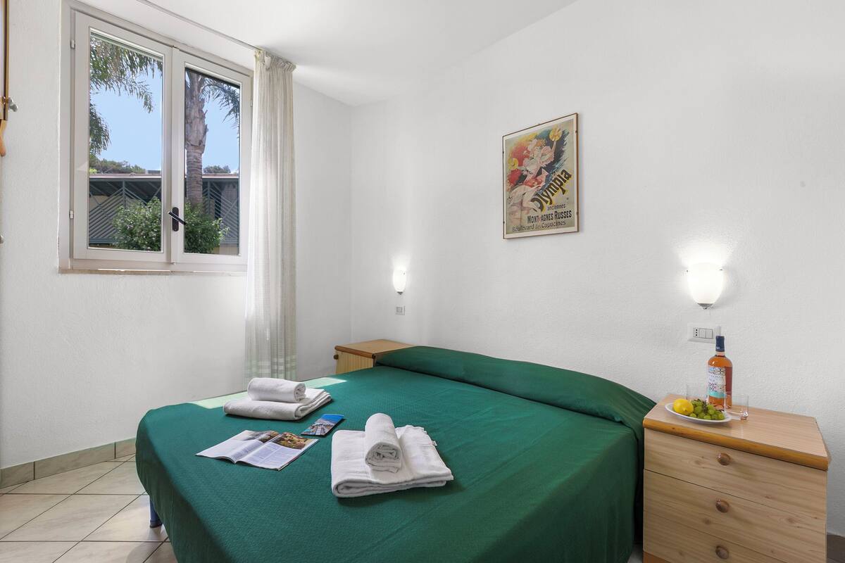 A hotel room with a bed, a nightstand with a bottle and glass, and a poster on the wall.