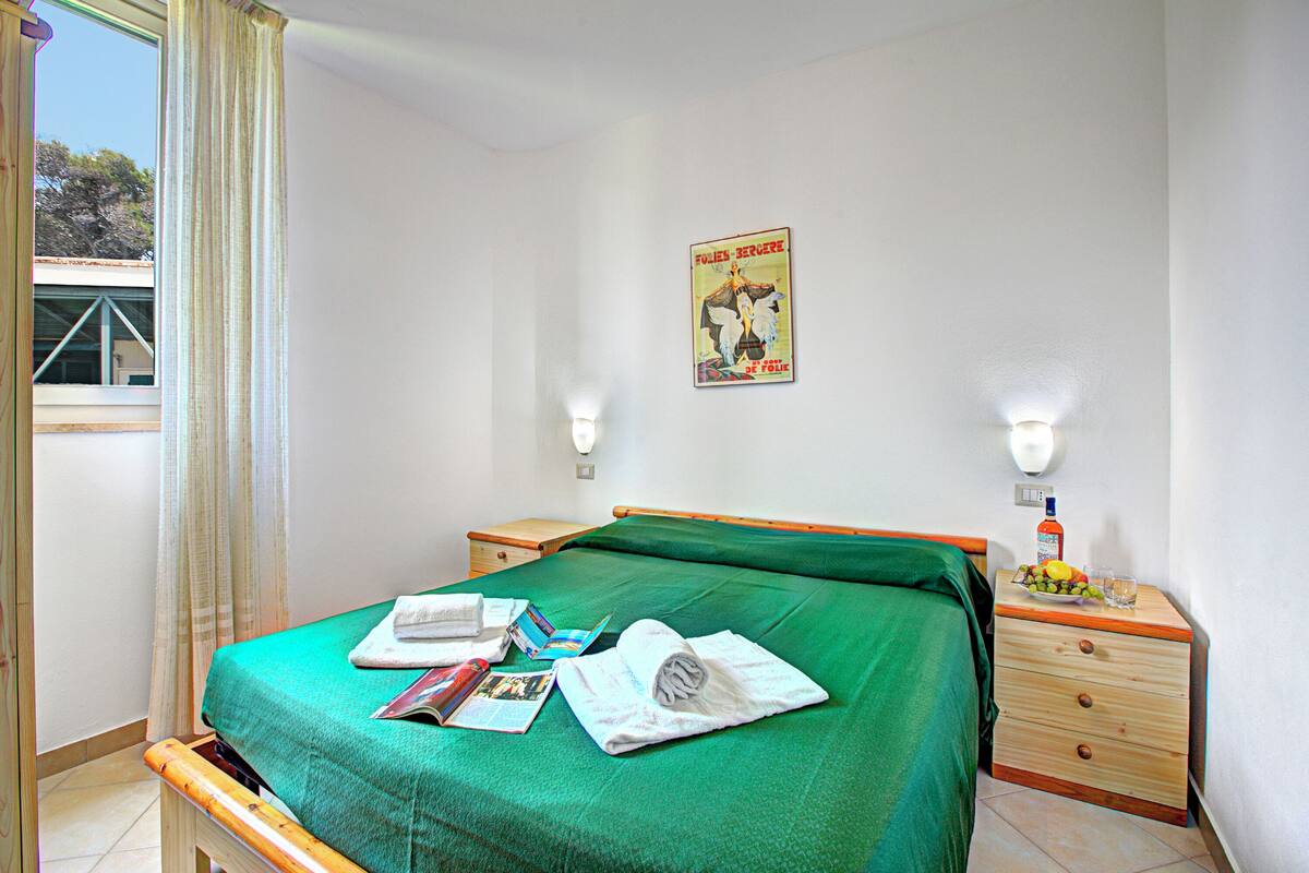 A hotel room with a bed, a nightstand, a poster on the wall, and a window with curtains.