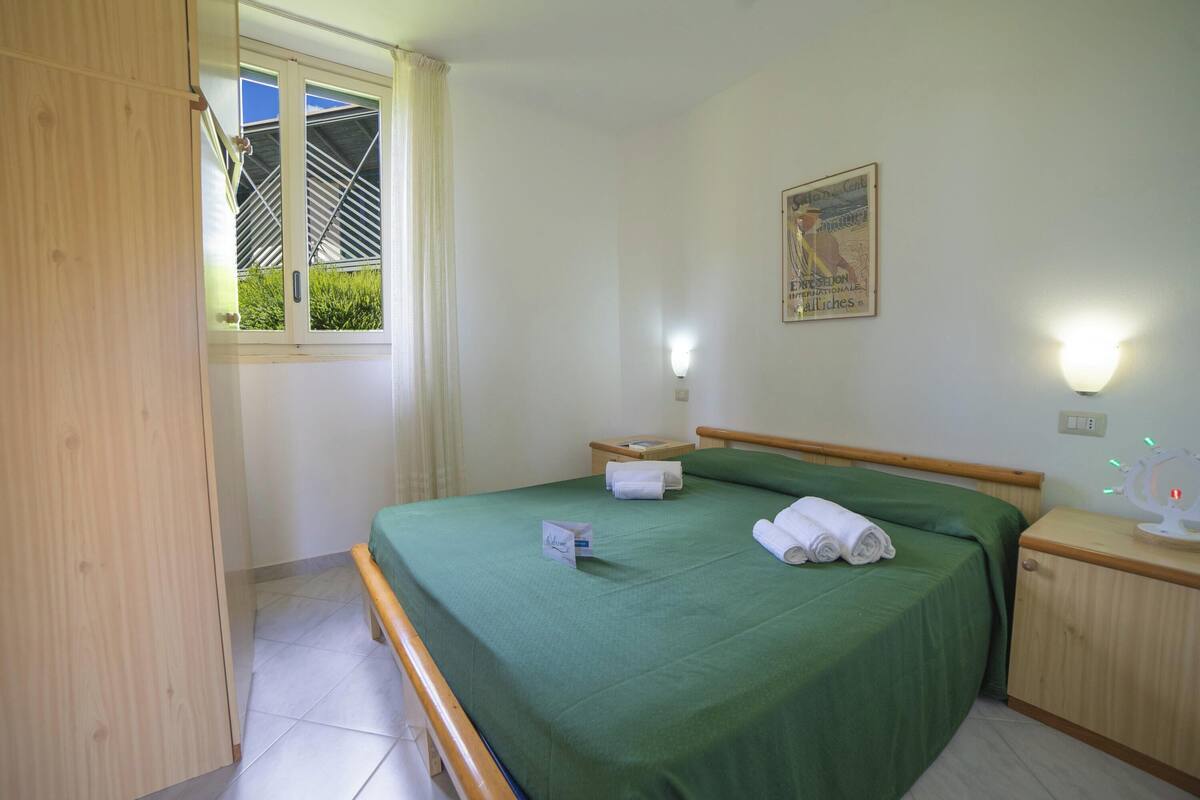 A hotel room with a single bed, a bedside table, a nightstand, a window with curtains, and a wall-mounted light fixture.