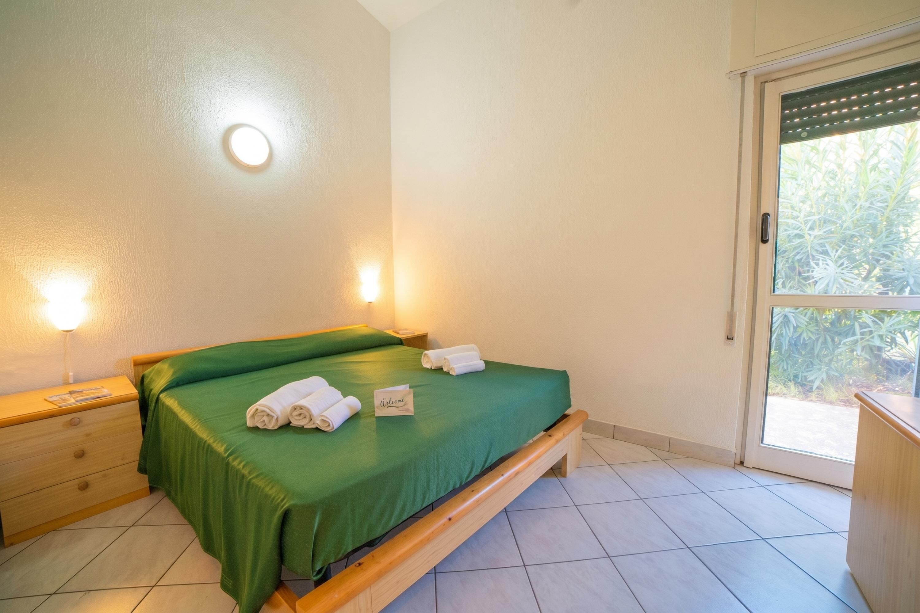 Ein Schlafzimmer mit grüner Bettdecke, einem Nachttisch mit Lampe und einem Fenster mit Blick ins Grüne.